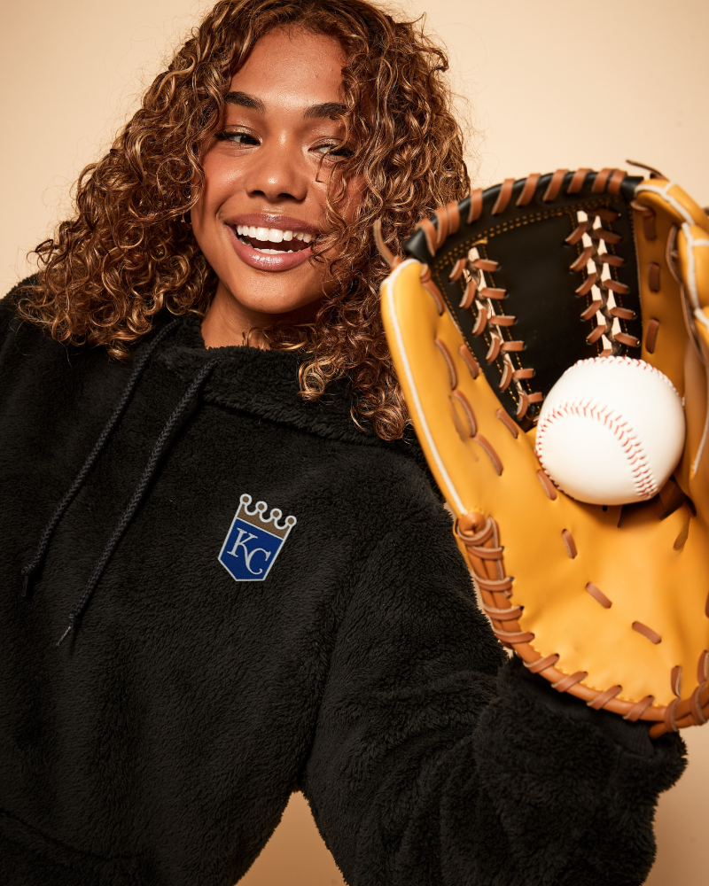 Person wearing a black hoodie with a kansas city royals logo, holding a baseball glove and ball against a beige background