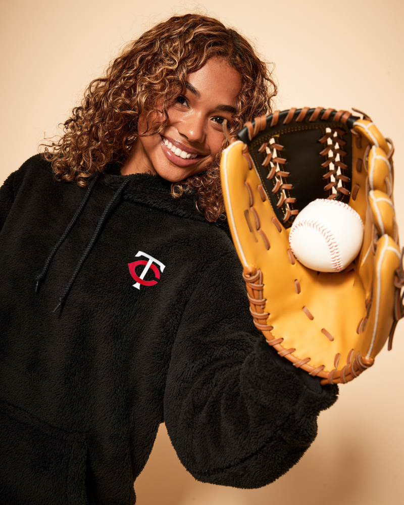 Person wearing a black hoodie with a minnesota twins logo, holding a baseball glove and ball against a beige background