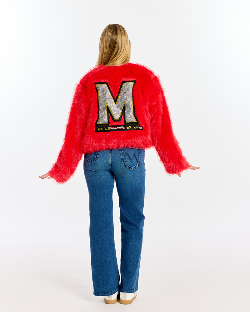 A woman viewed from behind wearing a bright red fuzzy cropped sweater with a large bedazzled silver 'M' patch on the back, paired with blue jeans and white shoes, against a plain white studio background.