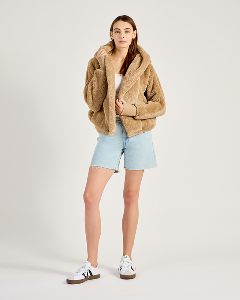 Person wearing a brown teddy coat, light blue shorts, and white sneakers on a plain background