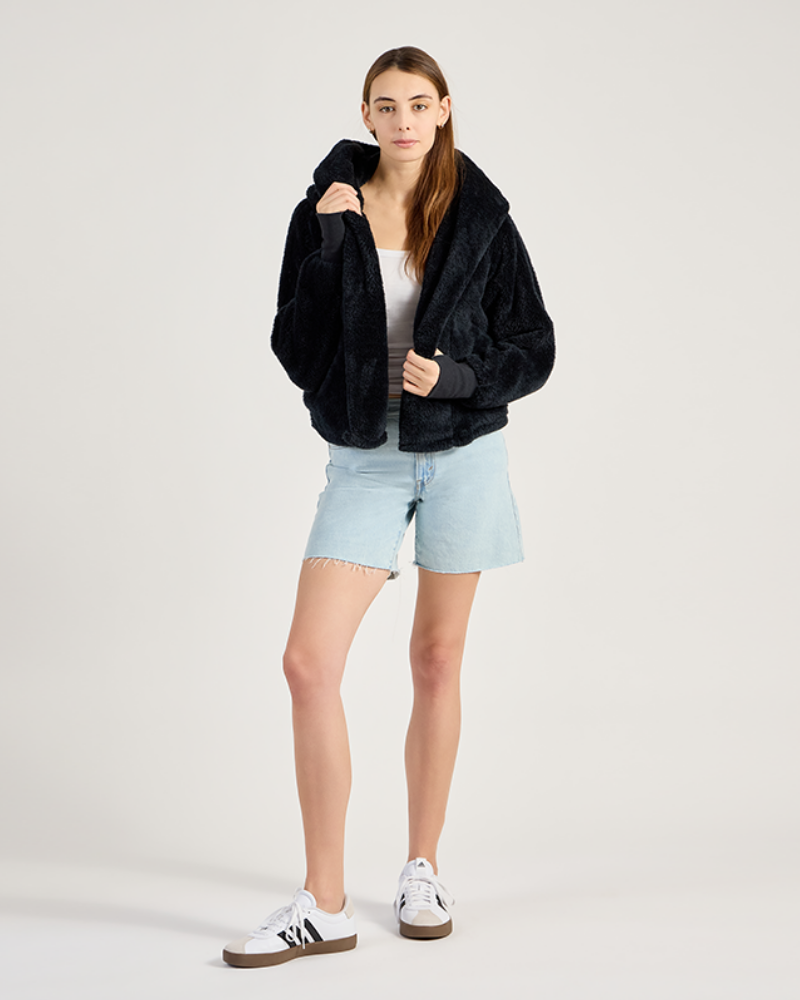 Person wearing a black jacket, light blue shorts, and white sneakers on a plain background