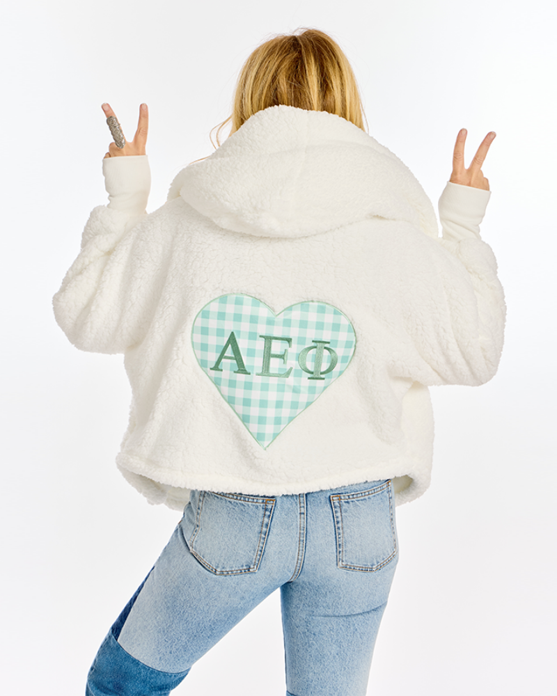 A person shown from the back wearing a white sherpa hoodie with a mint green and white gingham heart patch on the back featuring the Greek letters Alpha Epsilon Phi. The person is also wearing light blue jeans and posing with peace signs against a plain white background.
