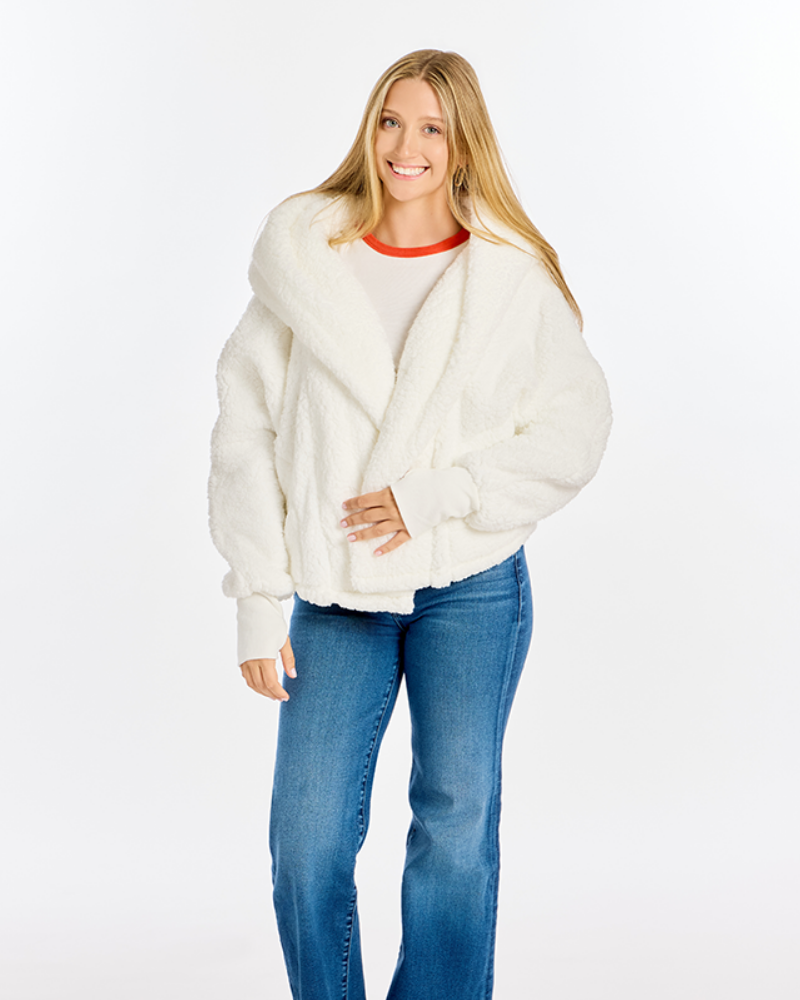 A woman modeling a fluffy off-white cropped jacket with a wide collar, worn over a white top with a red collar trim and blue jeans, against a plain white studio background.