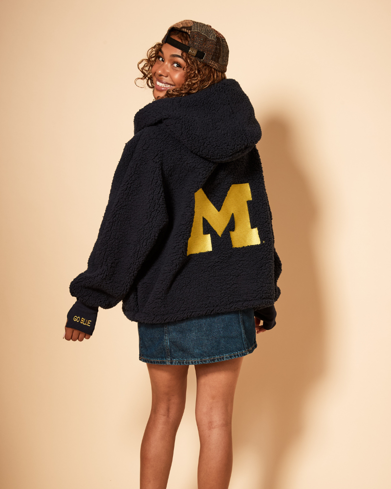 Person wearing a navy hoodie lounger with a large yellow university of michigan 'M' on the back against a beige background