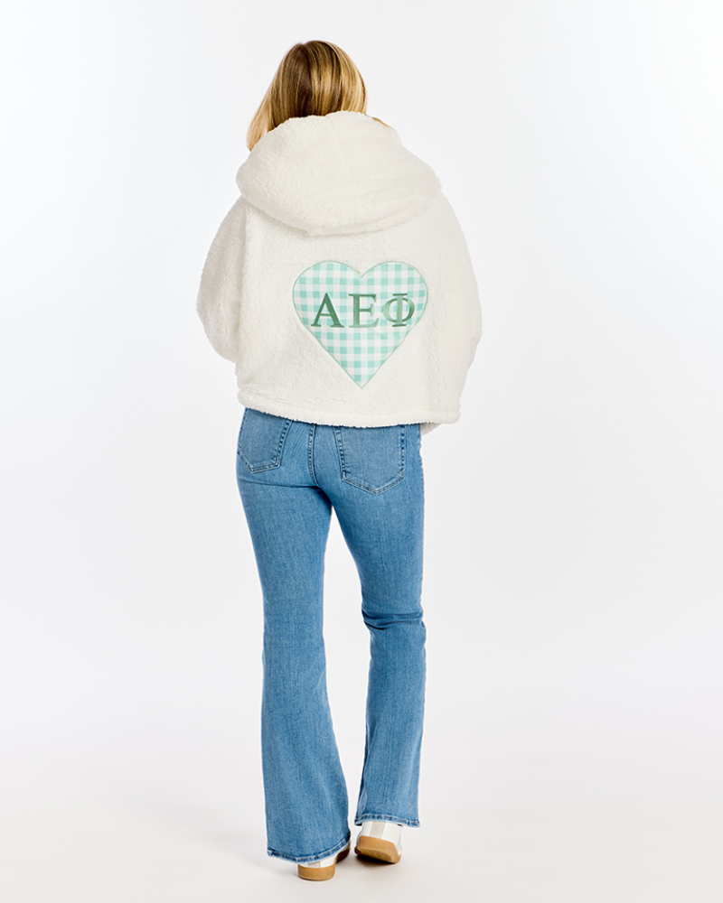 A woman viewed from behind wearing a cream-colored hooded sweatshirt with a mint green and white gingham heart patch on the back featuring the Greek letters AEPhi, paired with blue jeans, against a plain white studio background.