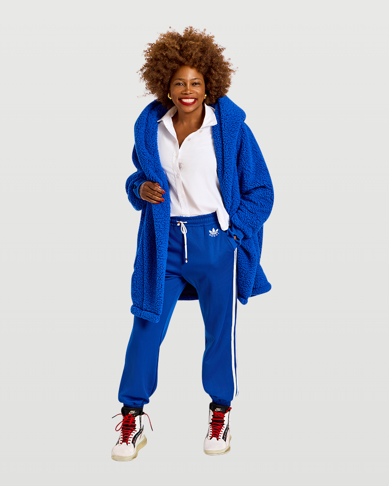 A woman modeling a matching blue tracksuit set consisting of a plush sherpa hooded cardigan and sweatpants with white Adidas stripes and logo, worn over a white collared shirt and paired with white sneakers.|Model is 5'7" wearing size XS/S