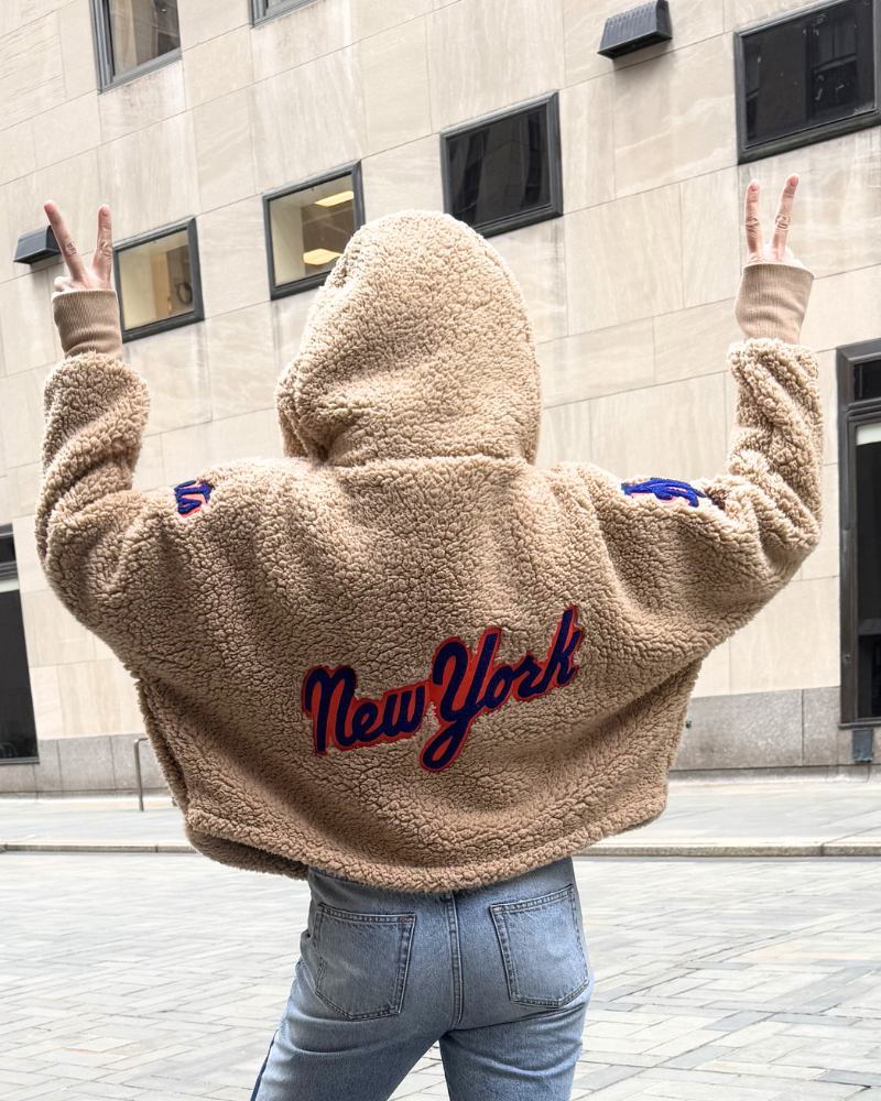 Person wearing a beige fleece jacket with 'New York' text, standing in front of a building.|Jill: 5'6" in size XS/S