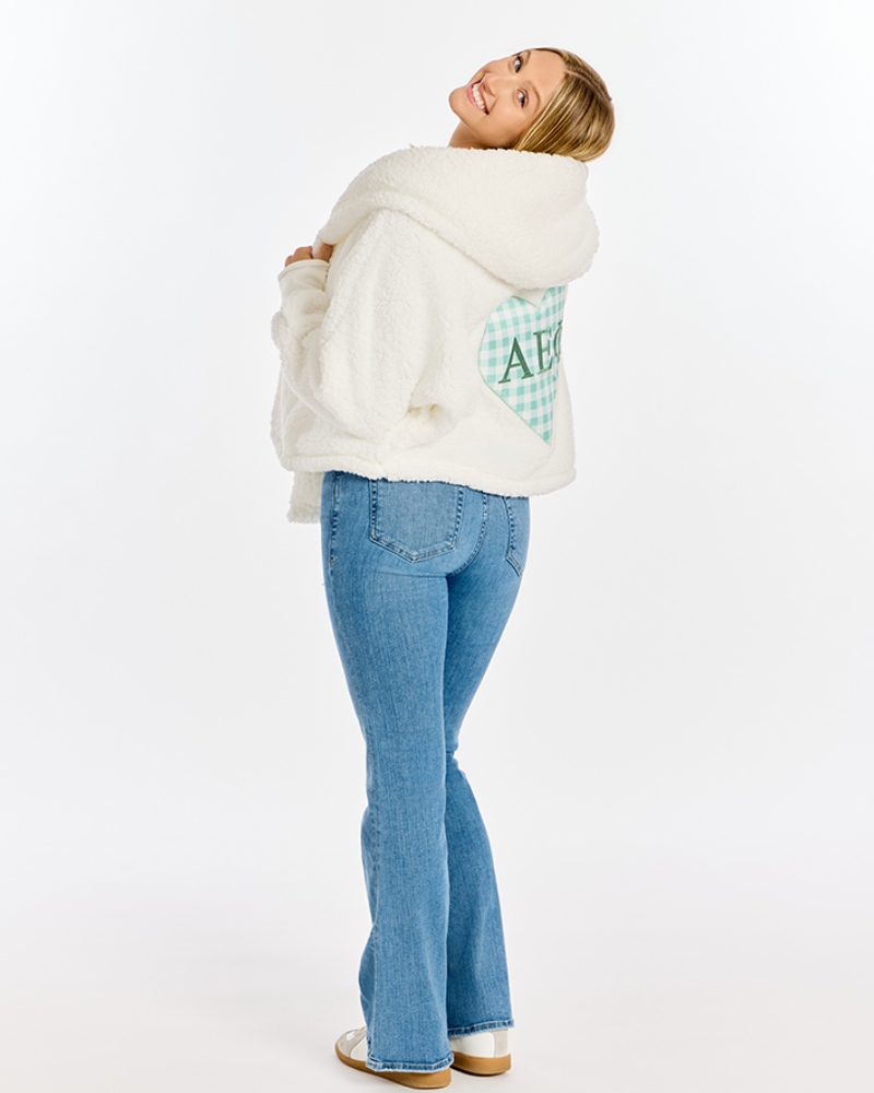 A woman wearing a white sherpa hoodie with a green and white checkered heart graphic on the back, paired with blue jeans and white sneakers, photographed against a plain white studio background.