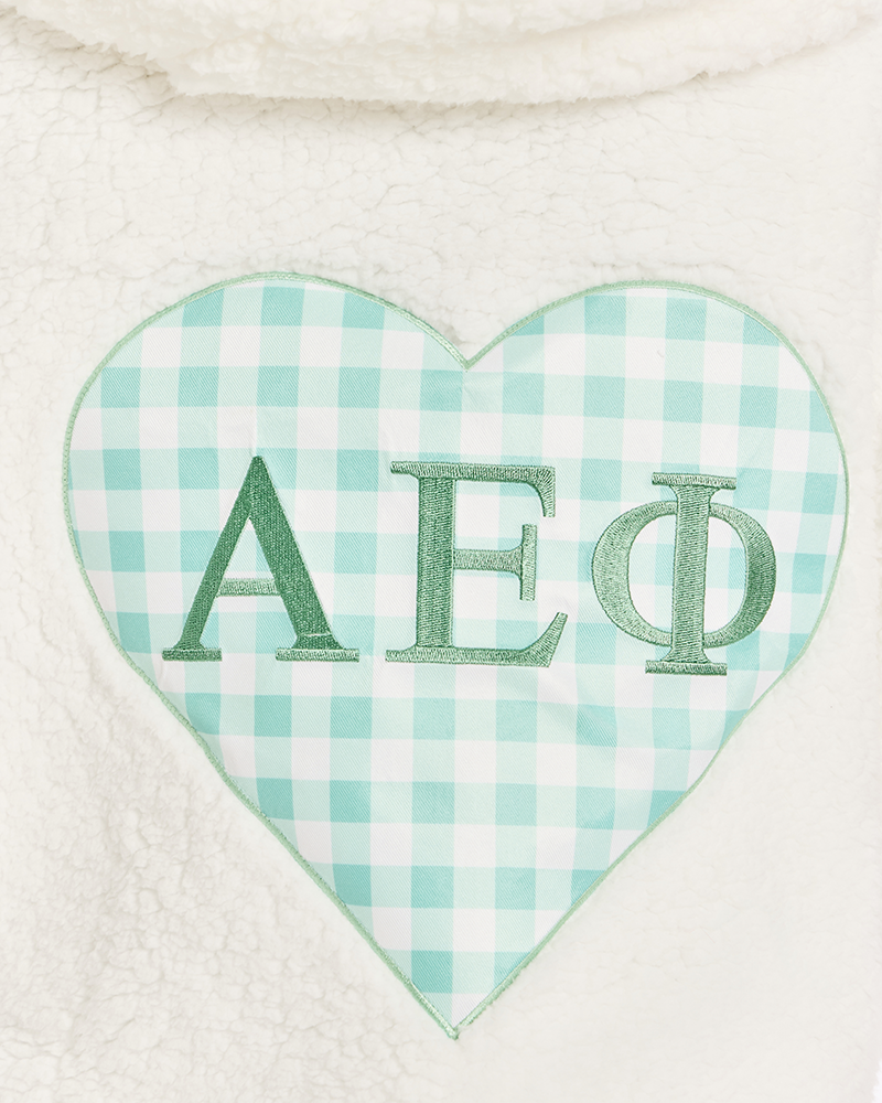 A close-up of a white sherpa fleece garment featuring a heart-shaped patch with a mint green and white gingham pattern and green embroidered Greek letters 'AEPhi' on it.