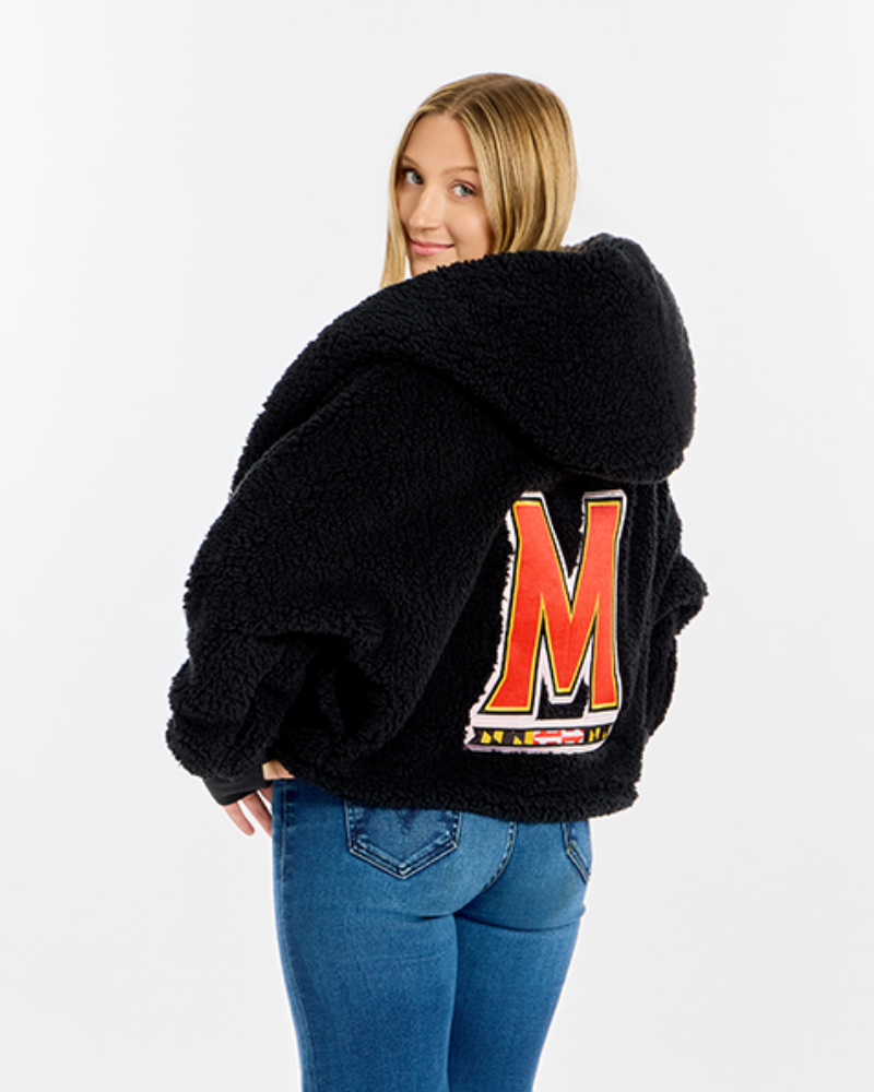 A woman wearing a black sherpa fleece hooded jacket with a large red and white 'M' patch on the back, paired with blue jeans, photographed against a light gray studio background.