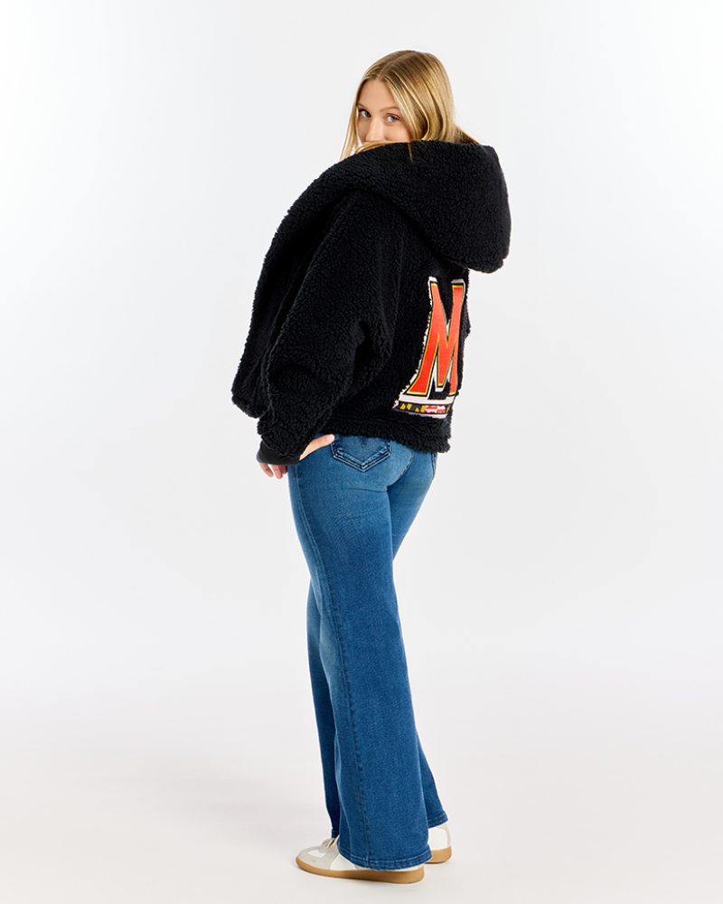 A woman wearing a black sherpa fleece cropped hoodie with a large red and white graphic 'M' on the back, paired with blue flare jeans and white sneakers, photographed against a seamless white studio background.