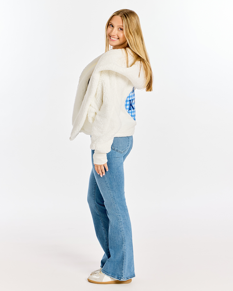 A young woman with blonde hair is shown from the back wearing a cream-colored fuzzy cropped sweater with a blue and white gingham pattern on the back, paired with blue flared jeans and white sneakers, against a plain white studio background.