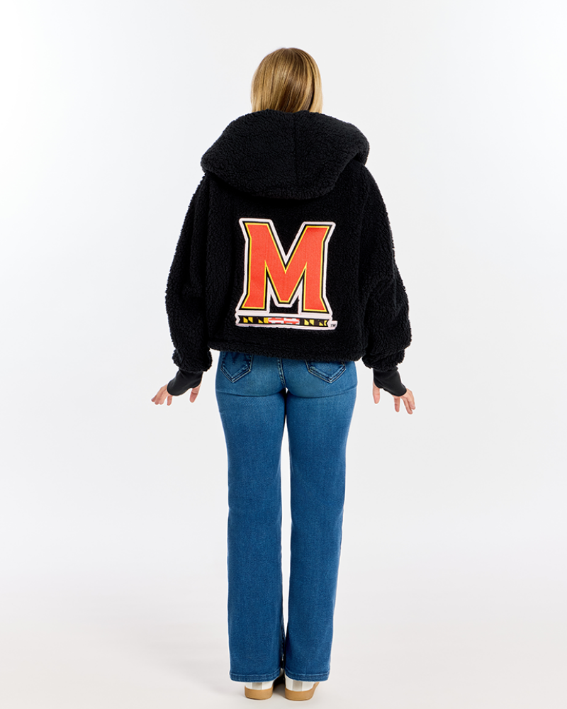 A woman viewed from behind wearing a black fuzzy hooded sweatshirt with a large red and white block letter 'M' on the back, paired with blue jeans, against a plain white studio background.