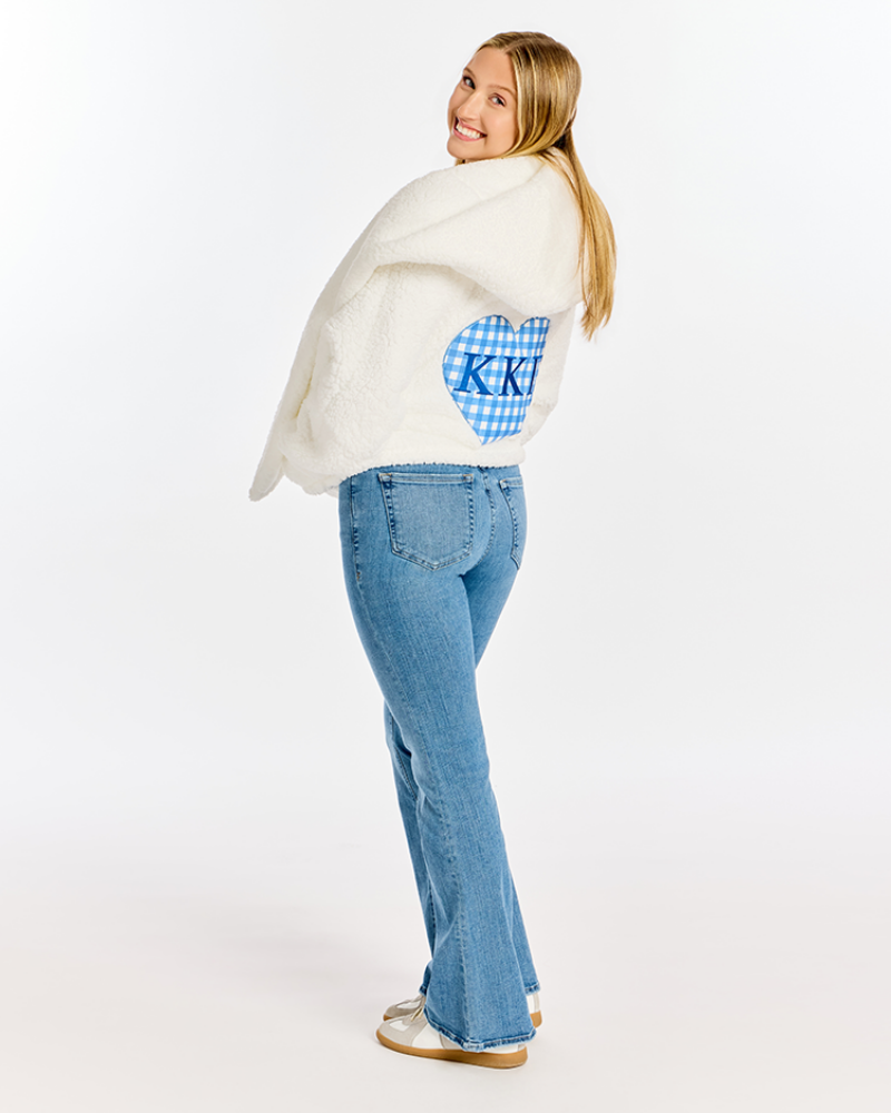 A woman wearing a white sherpa fleece jacket with a blue gingham heart patch on the back, paired with light blue jeans and white sneakers, photographed against a seamless white studio background.