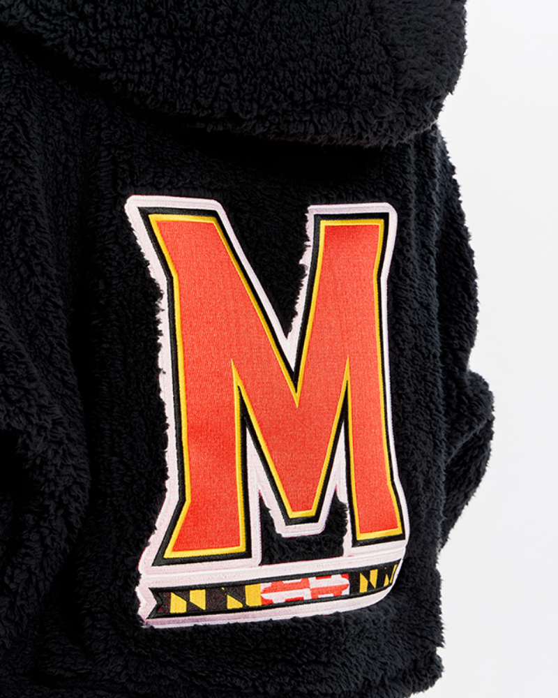 Close-up of a black sherpa fleece garment featuring a large embroidered letter 'M' patch with a white border, yellow inner border, and red fill, plus a small Maryland state flag detail below.