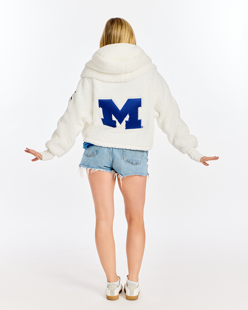 A woman viewed from behind wearing a cream sherpa hoodie with a large blue block letter 'M' on the back, paired with denim cutoff shorts and white sneakers, against a plain white studio background.