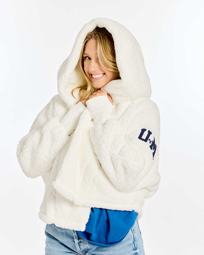 A woman wearing a fluffy white hooded sweatshirt with navy blue collegiate-style lettering on the sleeve, paired with blue jeans and a bright blue shirt underneath, against a plain white studio background.