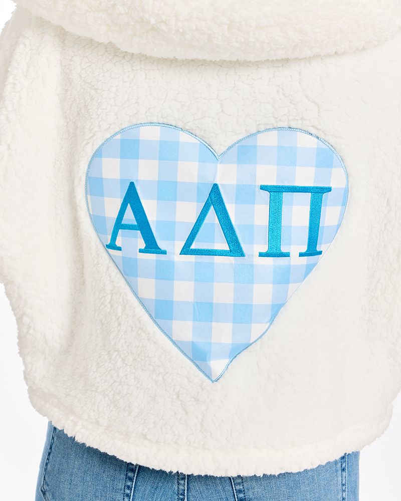 Close-up of a white sherpa fleece garment with a large heart-shaped patch featuring a light blue and white gingham pattern and the Greek letters Alpha Delta Pi in blue.
