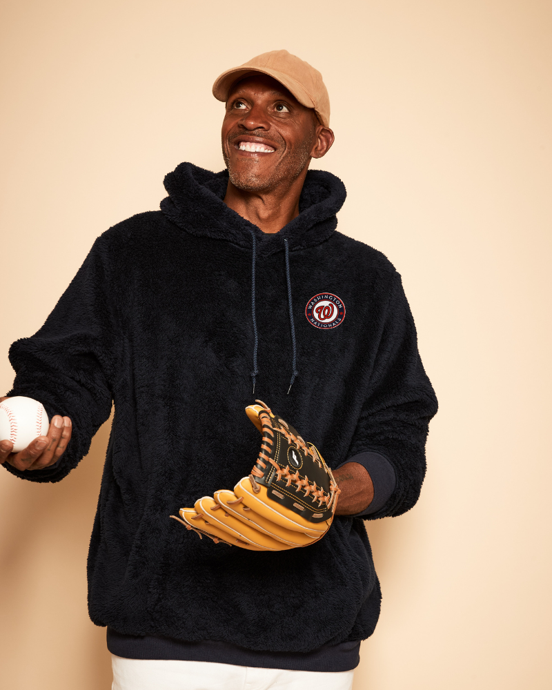 Man wearing a navy hoodie with a washington nationals logo, holding a baseball and glove against a beige background