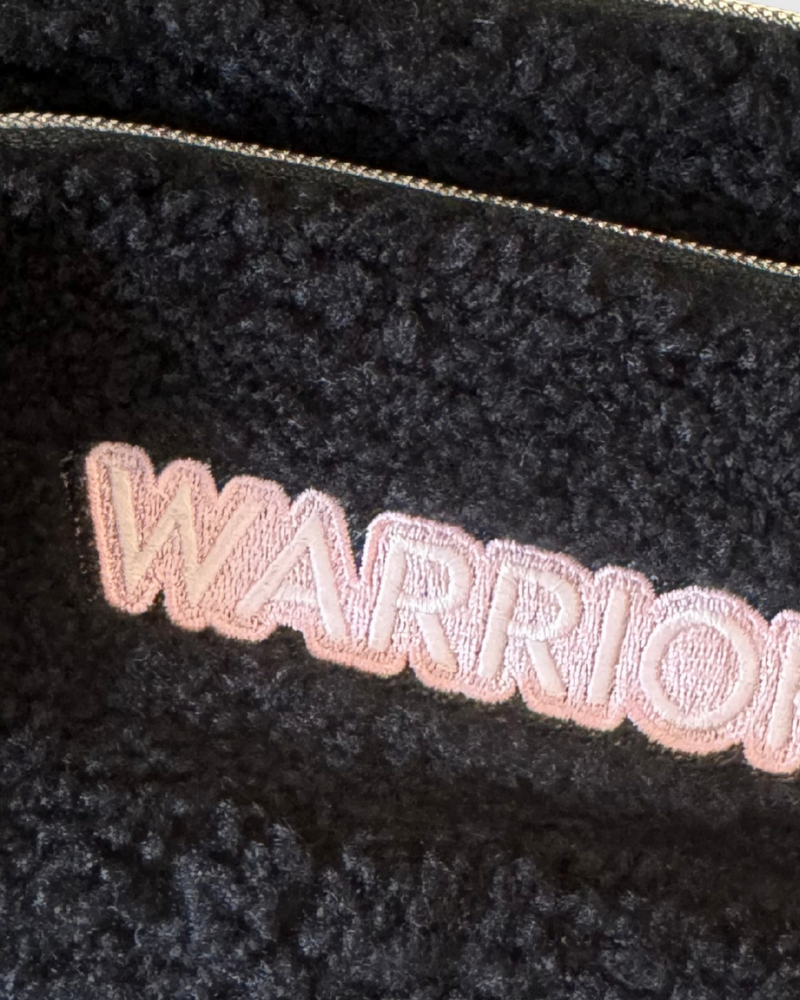 Close-up of a black sherpa or fleece pouch with a gold zipper and pink embroidered text reading 'WARRIOR'.