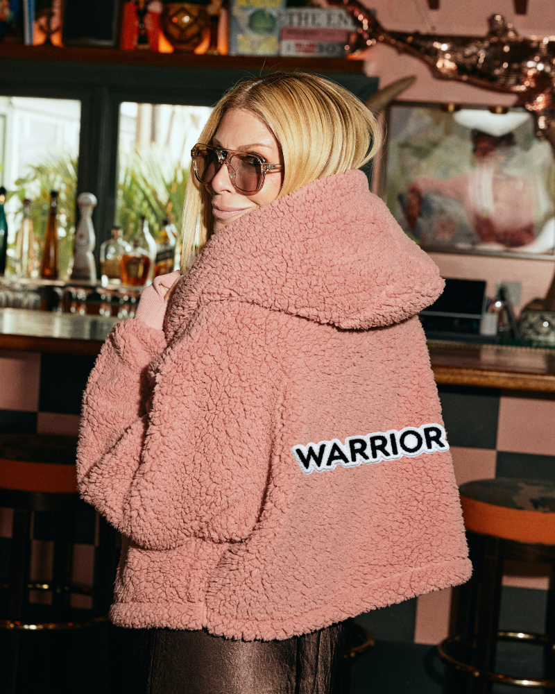 Person wearing a pink sherpa jacket with 'Warrior' logo in a bar setting