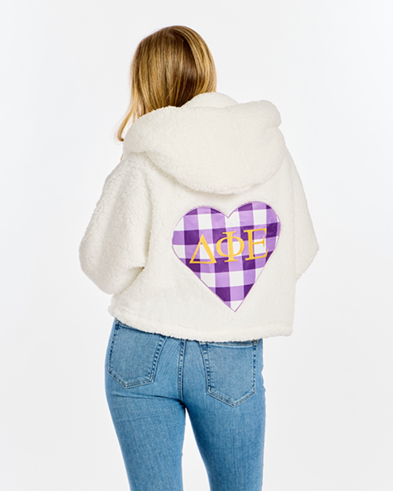 A woman viewed from behind wearing a cream sherpa fleece hooded jacket with a purple and white plaid heart graphic on the back featuring the Greek letters Delta Phi Epsilon in gold, paired with blue jeans against a white studio background.