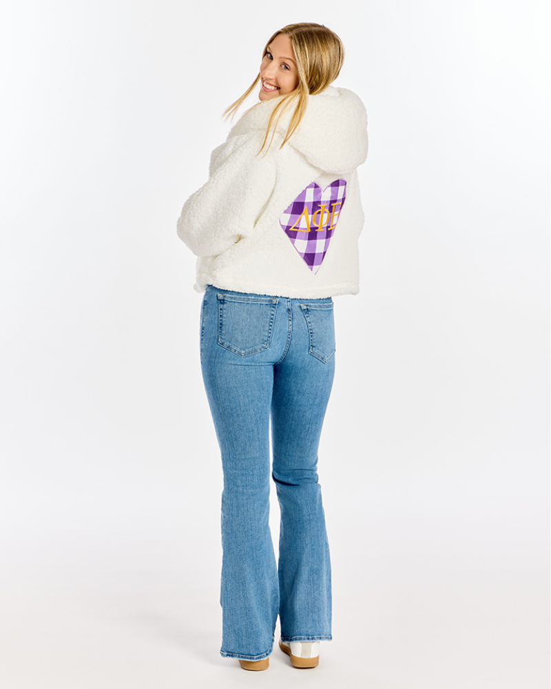 A woman wearing a white sherpa fleece hooded jacket with a purple and yellow plaid heart patch on the back, paired with blue jeans, photographed against a plain white studio background.