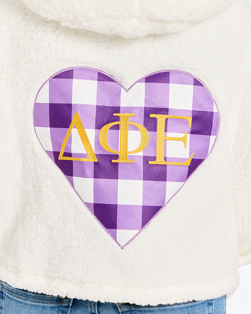 A close-up of a cream-colored sherpa hoodie featuring a large heart-shaped patch with a purple and white gingham pattern and gold Greek letters Delta Phi Epsilon.