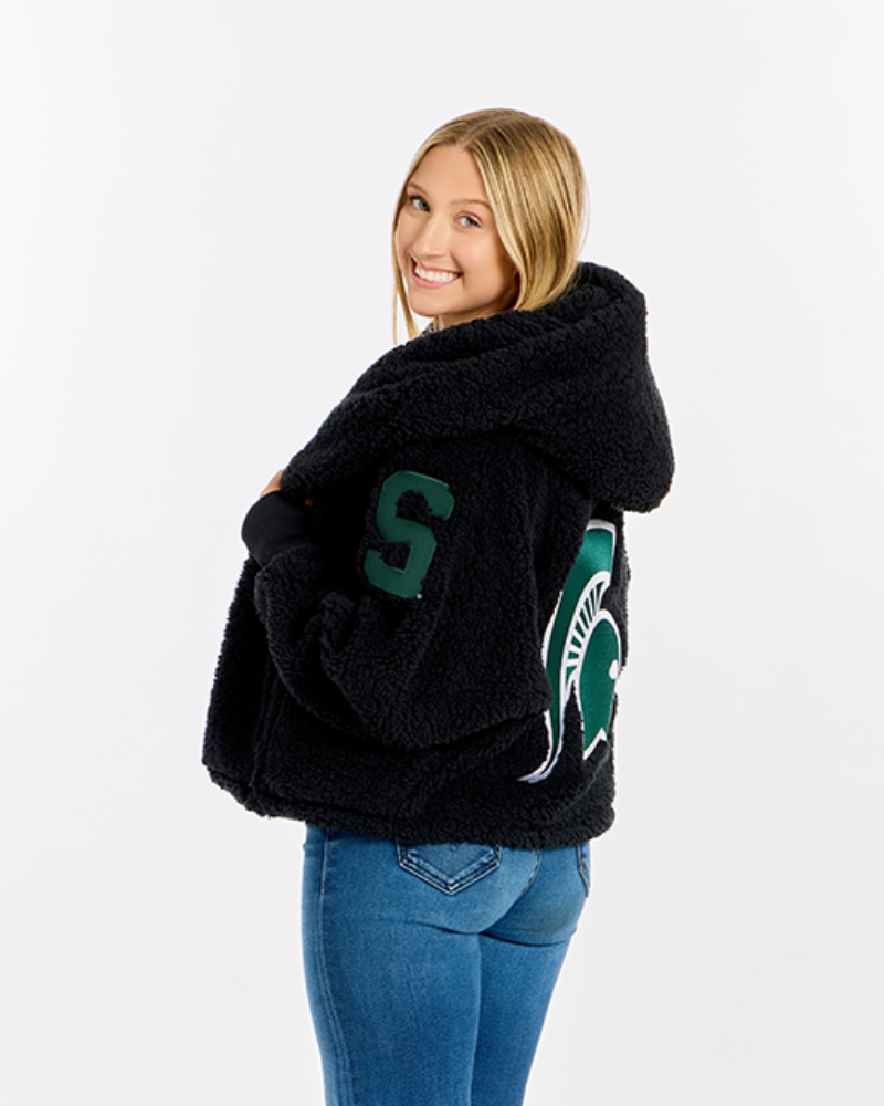 A woman modeling a black sherpa fleece hooded jacket with green and white Michigan State Spartans graphics, paired with blue jeans, against a plain white studio background.