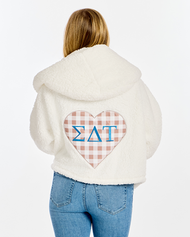 A woman viewed from behind wearing a cream sherpa hoodie with a large heart-shaped gingham patch on the back featuring the Greek letters Sigma Delta Tau in blue.