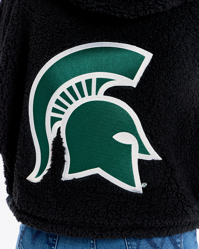 Close-up of a black sherpa hoodie featuring a large green and white Michigan State Spartans helmet logo on the front, worn with blue jeans.