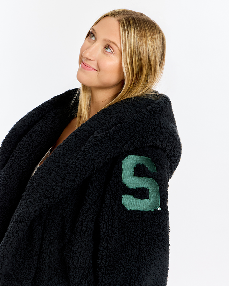A woman with blonde hair wearing a black sherpa fleece robe with a green letter 'S' patch on the sleeve, photographed against a solid white background.