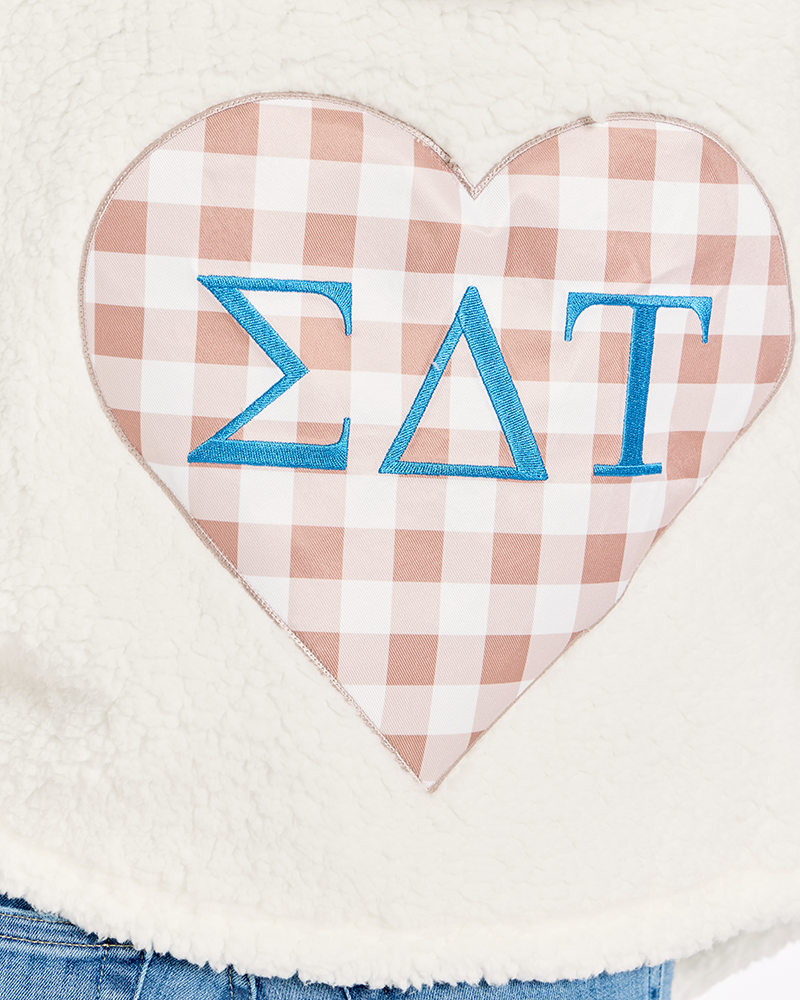 A close-up of a white sherpa fleece garment featuring a large heart-shaped patch with a pink and white gingham pattern and blue Greek letters 'Sigma Delta Tau' on it.