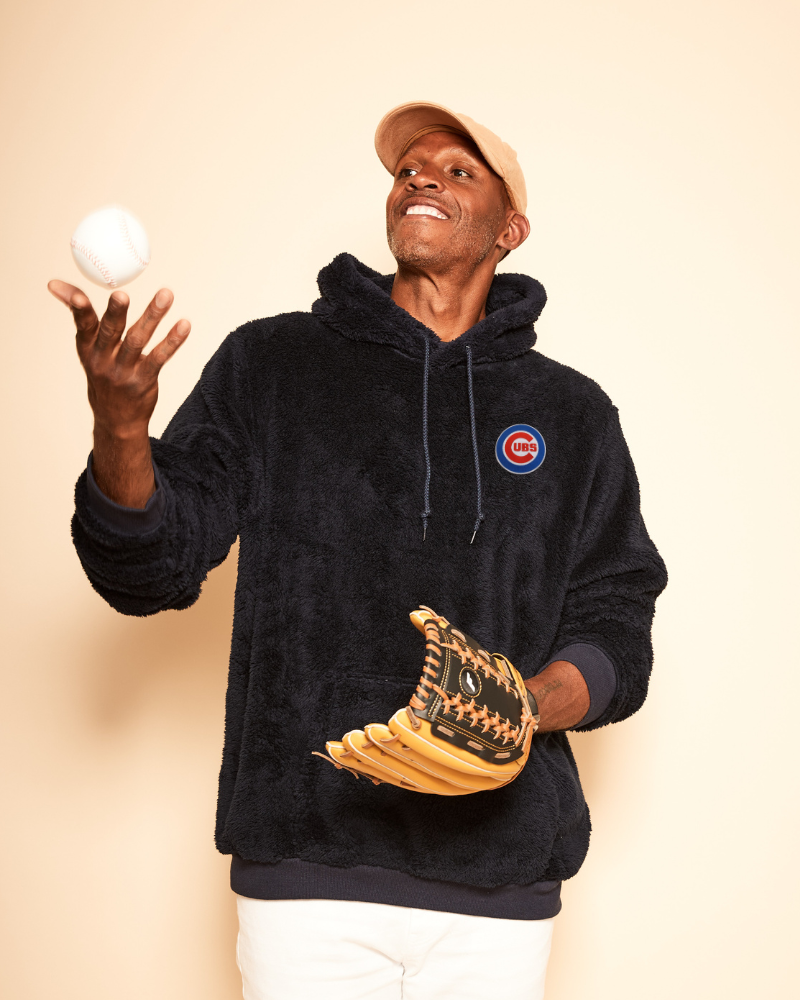 Person wearing a navy hoodie with a chicago cubs logo, holding a baseball and glove against a beige background