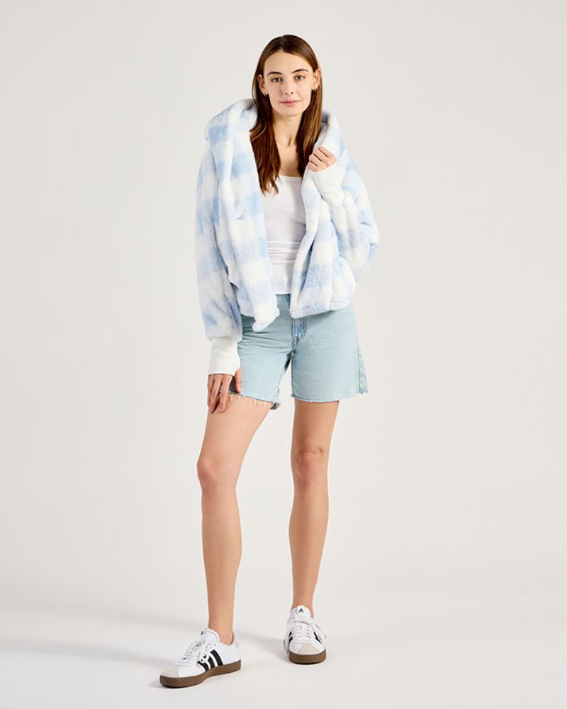 Woman wearing a gingham hoodie, white tank top, light blue shorts, and white sneakers on a plain background