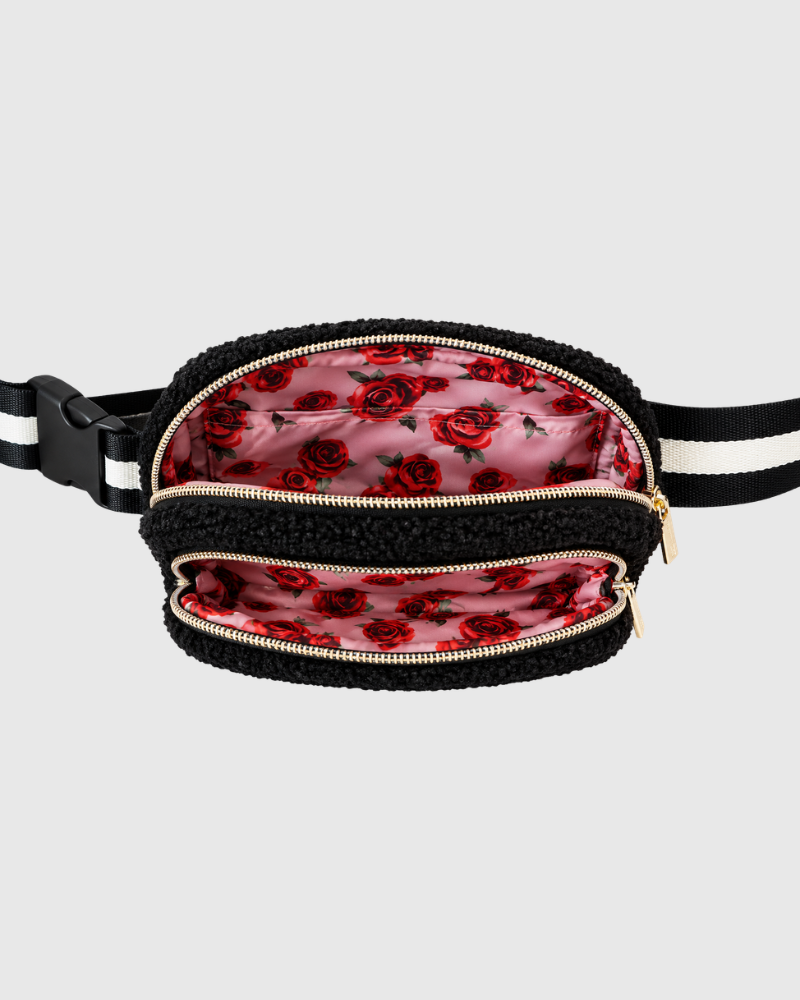 A black sherpa-lined waist bag with a black and white striped strap, shown open to reveal a pink satin lining printed with a red rose pattern. The bag has gold-tone zippers and is photographed against a solid light gray background.