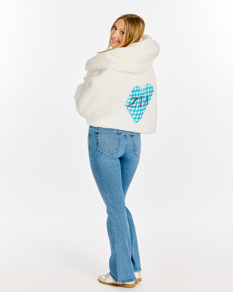 A woman wearing a white fuzzy cropped hoodie with a blue and white checkered heart patch on the back, paired with blue jeans and white sneakers, photographed against a plain white studio background.