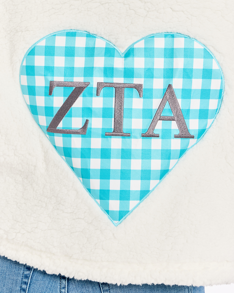 A close-up of a white sherpa fleece garment featuring a large turquoise and white gingham heart applique with the letters 'ZTA' in silver.