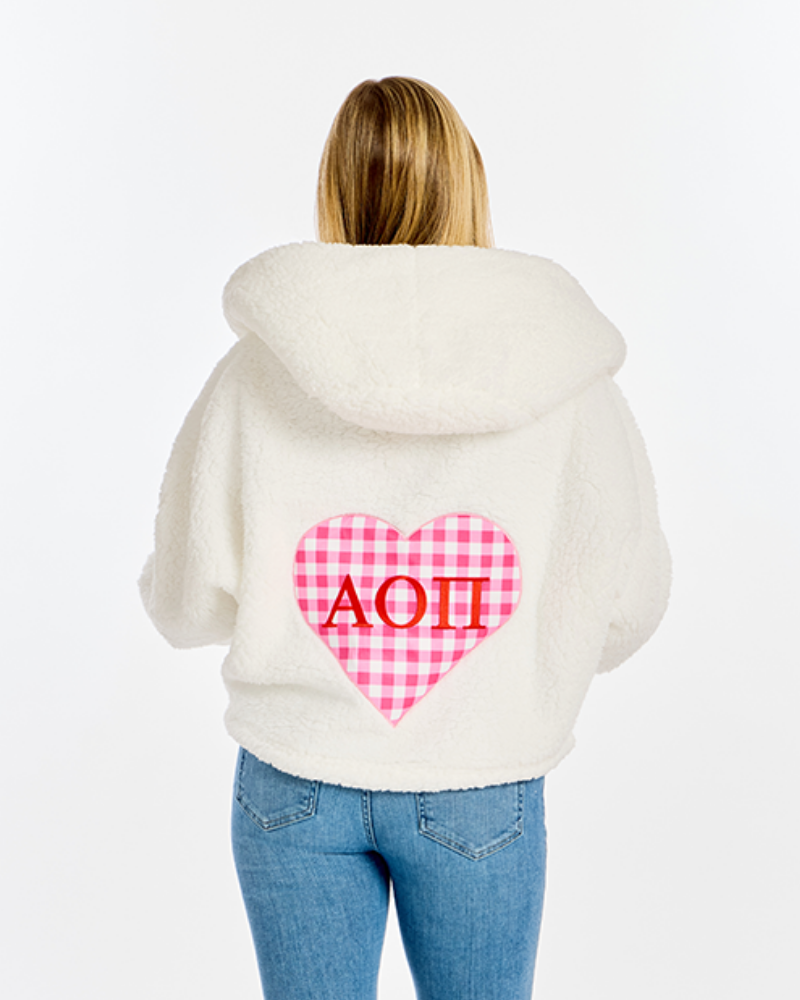 A woman viewed from behind wearing a cream sherpa hoodie with a pink and white gingham heart on the back featuring the red Greek letters 'ΑΟΠ', paired with blue jeans against a white studio background.