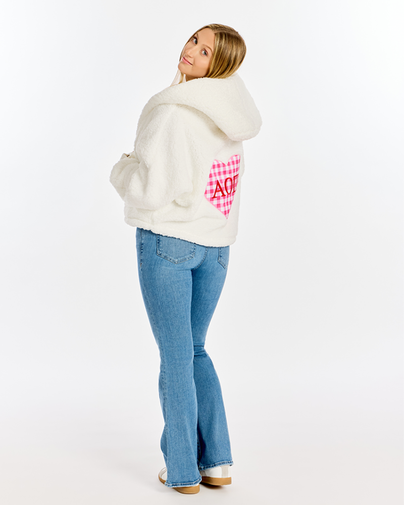 A woman wearing a white sherpa fleece hooded jacket with a pink and white gingham heart on the back, paired with blue jeans, photographed against a plain white studio background.