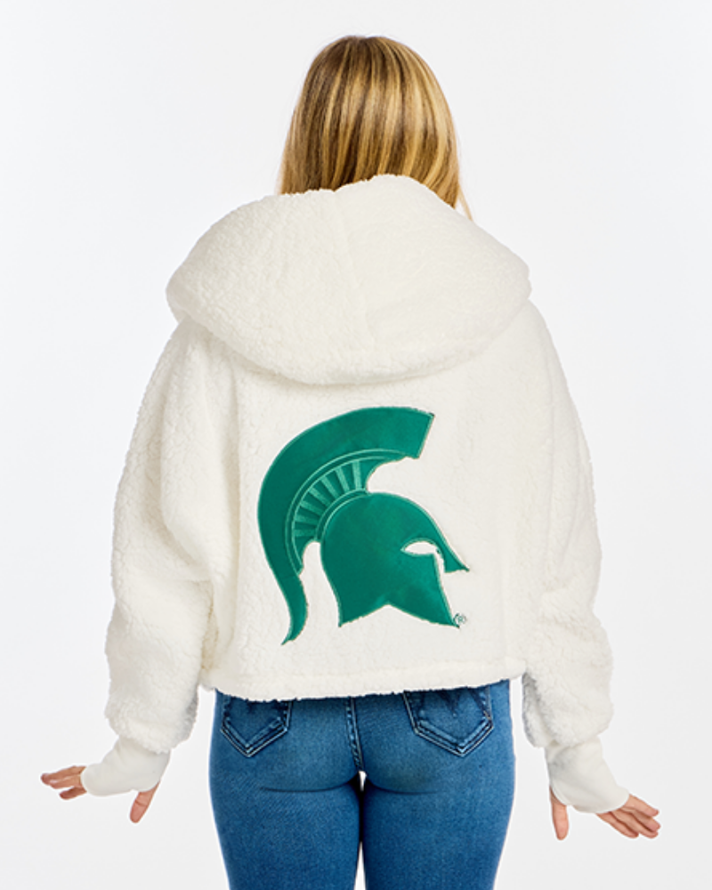 A woman viewed from behind wearing a white sherpa hoodie with a large green Michigan State Spartans helmet logo on the back, paired with blue jeans, against a plain white background.