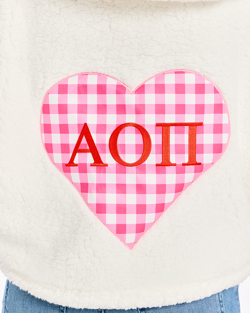 Close-up of a cream sherpa fleece garment with a large pink and white gingham heart patch on the front featuring red Greek letters 'Alpha Omicron Pi'.