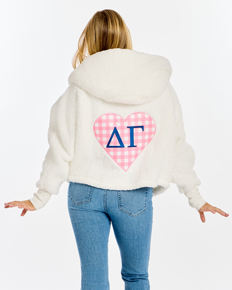 A woman viewed from behind wearing a cream-colored fuzzy cropped hoodie with a pink and white gingham heart graphic on the back featuring blue Greek letters Delta Gamma, paired with blue jeans, against a plain white background.