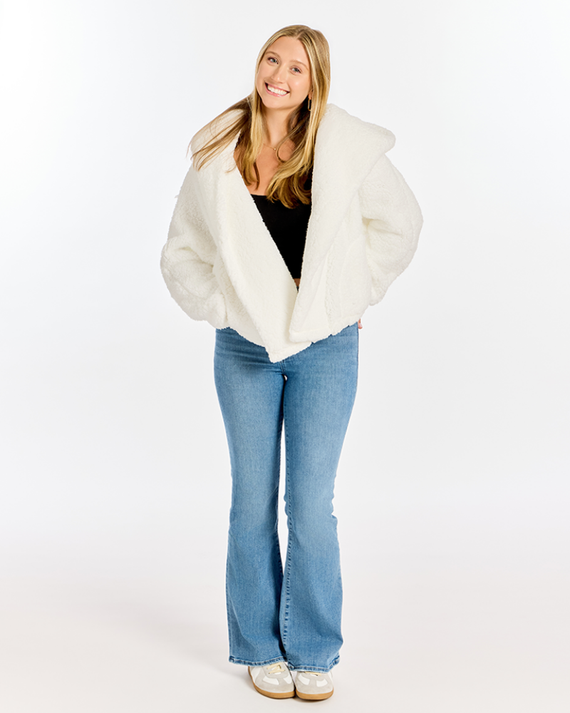 A woman modeling a fluffy off-white faux fur jacket with a wide collar, worn over a black top and blue flare jeans, against a plain white studio background.