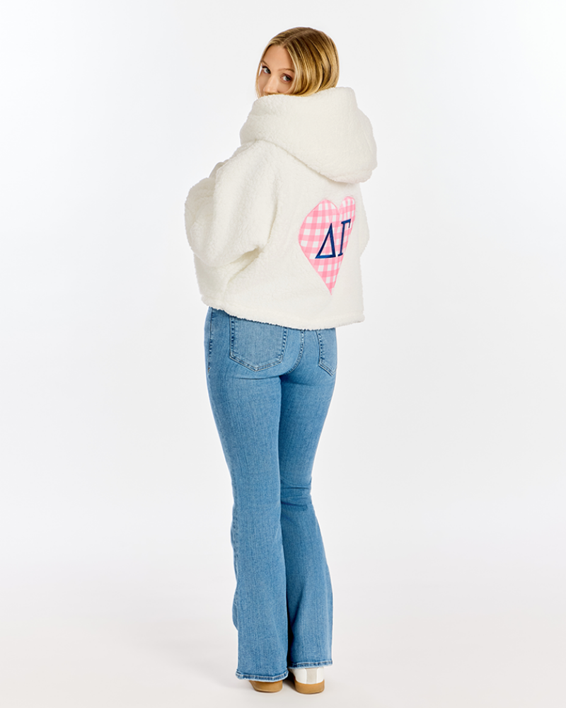 A woman wearing a cream sherpa hoodie with a pink and white gingham heart and a blue Delta Gamma symbol on the back, paired with blue jeans, photographed from behind against a white studio background.