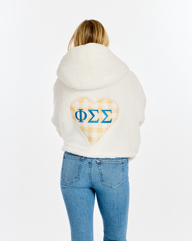 A woman viewed from behind wearing a cream sherpa hoodie with a yellow-and-white gingham heart graphic and blue Greek letters on the back, paired with blue jeans, against a plain white background.