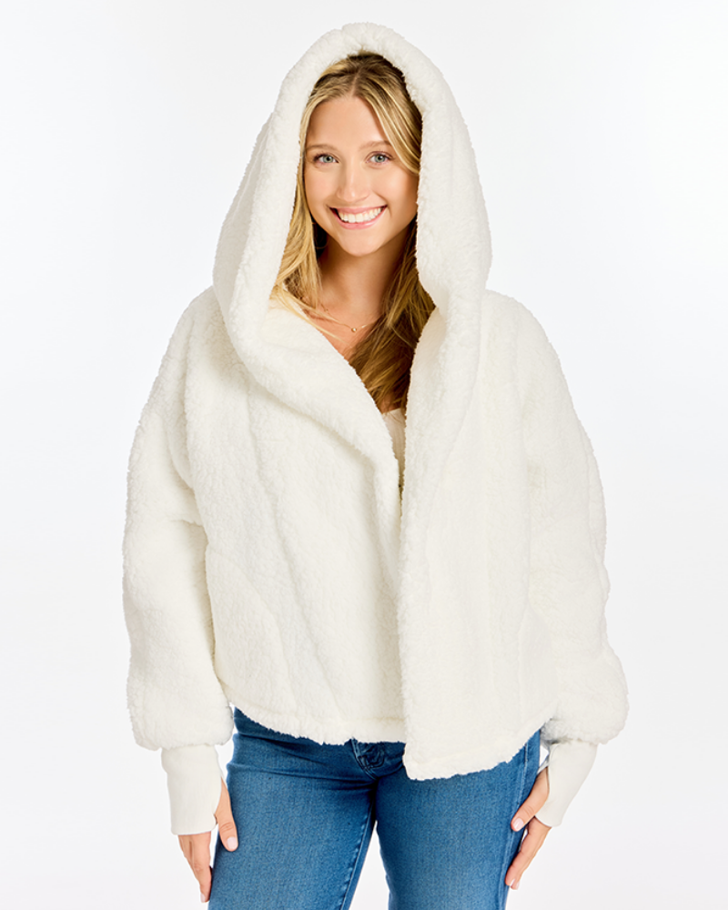 A smiling blonde woman wearing a fluffy white hooded jacket with a shawl collar and ribbed cuffs, paired with blue jeans, against a plain white studio background.