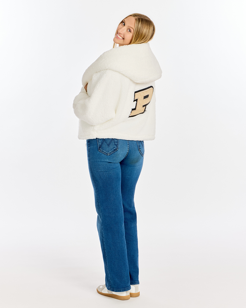 A woman wearing a white sherpa fleece hooded jacket with a large beige and black embroidered letter 'P' on the back, paired with blue jeans and white sneakers, photographed against a seamless white studio background.