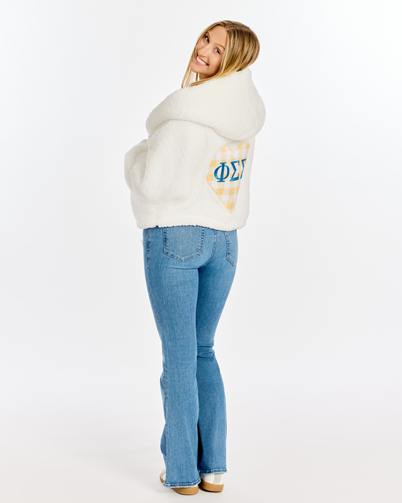 A woman wearing a fluffy white sherpa hoodie with a yellow and white plaid patch featuring blue Greek letters on the back, paired with blue jeans, photographed against a solid white studio background.
