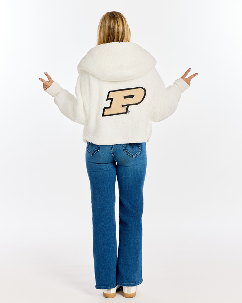 A woman viewed from behind wearing a white sherpa hoodie with a large tan and black 'P' logo on the back, paired with blue jeans, against a plain white studio background.